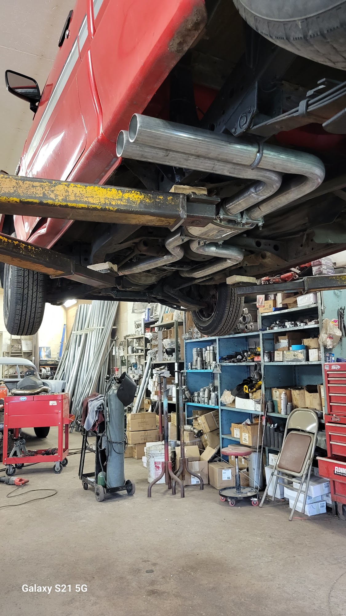 Classic red truck on a lift with custom exhaust routing visible, parts shelves in background
